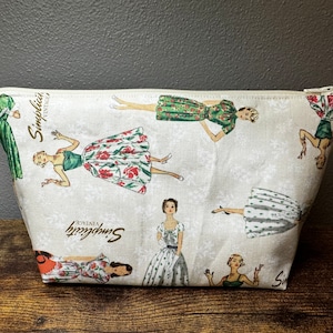 May include: A white zippered pouch with a vintage Simplicity pattern featuring women in various dresses. The pattern includes red, green and white floral prints. The pouch has the text "Simplicity Vintage" printed on it.