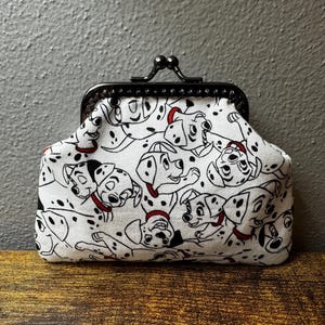 May include: A white coin purse featuring a repeating pattern of black and white Dalmatian puppies with red collars. The pouch has a black metal kiss-lock frame closure at the top, providing a secure way to store small items inside the fabric container.