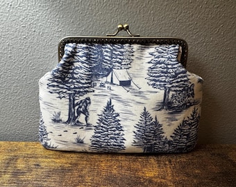 May include: A fabric coin purse featuring a dark blue and cream toile-style print of a forest scene with pine trees, a tent, and a Bigfoot figure. The pouch has a metallic bronze-toned kiss-lock frame closure at the top with a hinged mechanism.