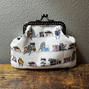 May include: A white fabric coin purse with a black metal clasp. The purse is patterned with gray cats reading books on shelves. The books are in various colors, and the background is a light gray.