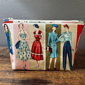 May include: A white zippered pouch with a colourful print of five women in vintage dresses. The women are wearing blue, red, green, and white dresses with polka dots and floral patterns.