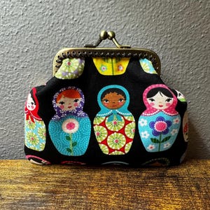 May include: A black fabric coin purse with a metal clasp, featuring a pattern of colorful Matryoshka dolls. The dolls have different skin tones and floral designs on their clothing. The purse is on a wooden surface.