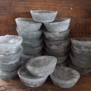 May include: A collection of small, translucent, gray-green bowls stacked in multiple piles. The bowls have a rounded shape with a slightly rough texture, and are arranged against a wooden background.