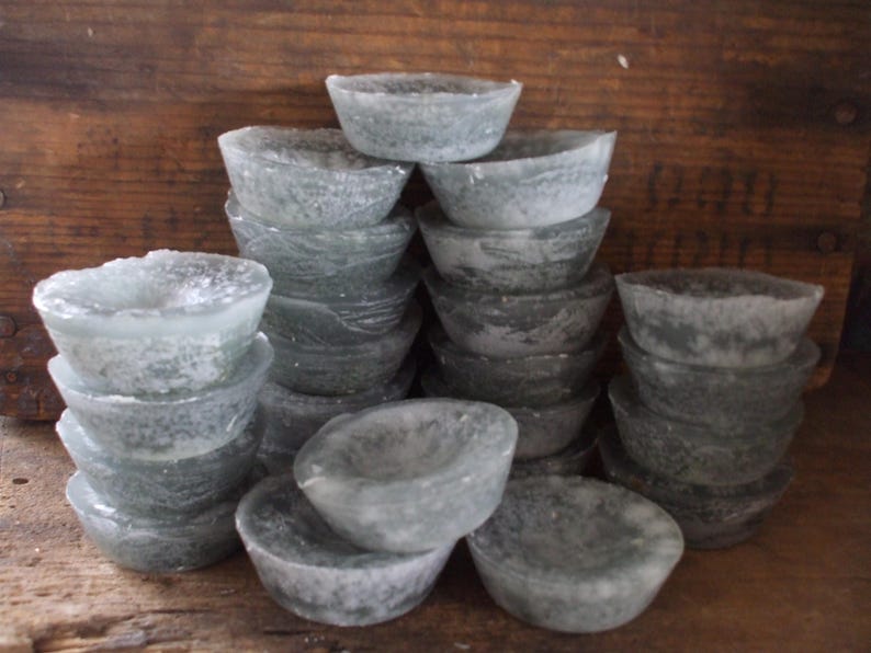 May include: A collection of small, translucent, gray-green bowls stacked in multiple piles. The bowls have a rounded shape with a slightly rough texture, and are arranged against a wooden background.