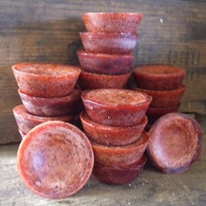 May include: A stack of 10 small, red, round, wax candle holders. The candle holders are stacked in a pyramid shape. The candle holders are made of a red wax with a textured surface.