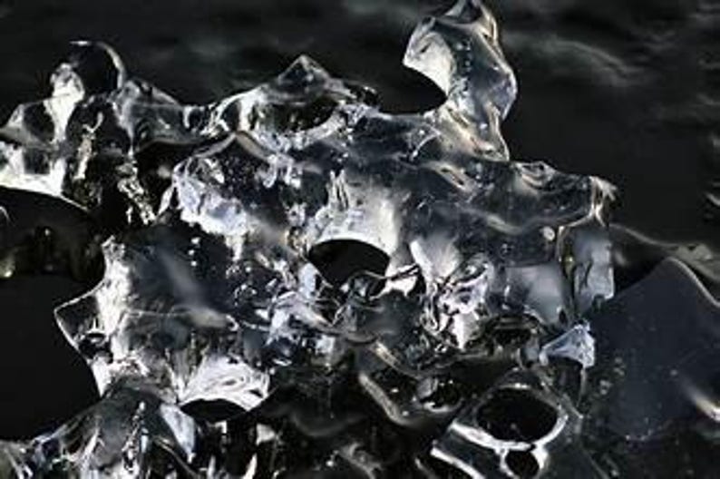 May include: Close-up of a translucent, irregularly shaped ice formation. The ice has a complex, textured surface with multiple peaks and valleys, reflecting light and creating a sparkling effect. The background is dark, providing contrast to the ice.