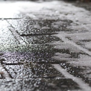 May include: Close-up of a wet, dark gray brick walkway with patches of snow and ice. The bricks are rectangular and arranged in a grid pattern. The surface reflects light, creating a shiny effect.