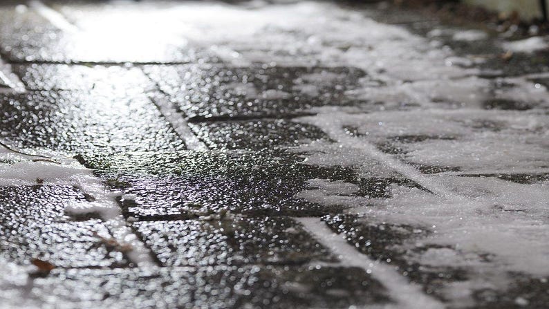 May include: Close-up of a wet, dark gray brick walkway with patches of snow and ice. The bricks are rectangular and arranged in a grid pattern. The surface reflects light, creating a shiny effect.