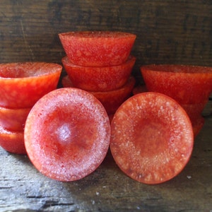 May include: A stack of eight red, round, wax melts with a frosted finish. The wax melts are arranged in a pyramid shape on a wooden surface.