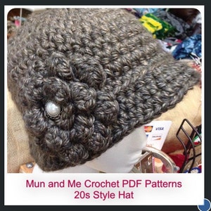 Puede incluir: Un gorro de crochet gris con un detalle de flor y un acento de perla. El gorro tiene una visera y está diseñado en un estilo de los años 20. Patrones de crochet PDF Mun and Me Gorro estilo años 20.
