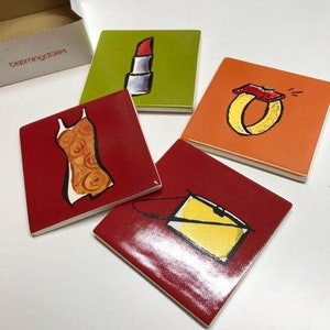 May include: Four square ceramic coasters with colorful illustrations of a red lipstick, a gold ring, a yellow purse, and a red swimsuit. The coasters are all red on the back and have the Bloomingdale's logo on the box.