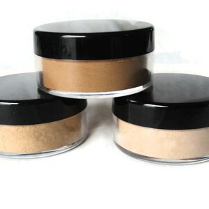 May include: Three round, clear plastic cosmetic jars with black screw-on lids, each filled with a fine, loose powder in varying shades of beige and tan, arranged in a stacked formation against a plain white background.