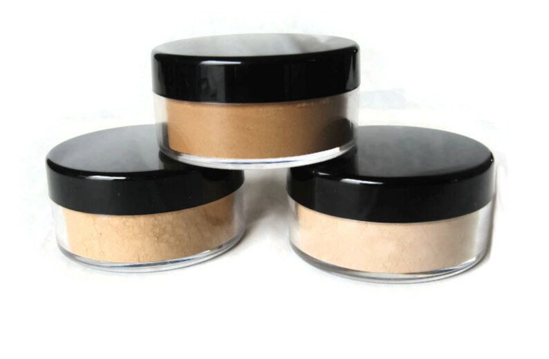 May include: Three round, clear plastic cosmetic jars with black screw-on lids, each filled with a fine, loose powder in varying shades of beige and tan, arranged in a stacked formation against a plain white background.