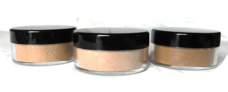 May include: Three round, clear plastic cosmetic jars with black screw-on lids, each containing a different shade of loose face powder ranging from light beige to medium tan, arranged in a horizontal row against a plain white background.