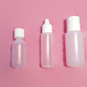 May include: Three clear plastic bottles with white caps on a pink background. The bottles are different sizes. The smallest bottle is on the left, the middle bottle is in the center, and the largest bottle is on the right.