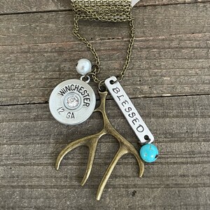 May include: A necklace with a silver bullet charm that says "Winchester 12 GA", a small antler charm, a silver charm that says "Blessed", a white pearl bead, and a turquoise bead. The necklace is on a brown wooden surface.