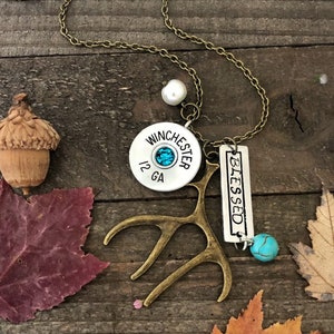 May include: A necklace with a silver charm that says "Blessed" and a Winchester 12 GA shotgun shell charm. The necklace also has a small antler charm and a turquoise stone charm.