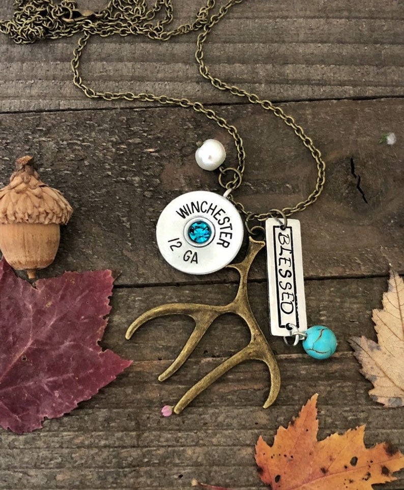 May include: A necklace with a silver charm that says "Blessed" and a Winchester 12 GA shotgun shell charm. The necklace also has a small antler charm and a turquoise stone charm.
