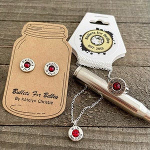 May include: A set of silver bullet earrings, a necklace, and a ring. The earrings and necklace feature red rhinestones and the text "WINCHESTER 38 SPL+P" and "WINCHESTER 9mm LUGER". The ring features a red rhinestone and the text "WINCHESTER AUTO". The set is packaged on a brown card with the text "Bullets For Belles By Katelyn Christie".