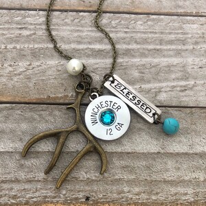 May include: A silver necklace with a variety of charms, including a shotgun shell with a turquoise stone, a small antler, a pearl, and a silver tag that says "Blessed".