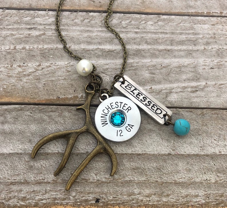 May include: A silver necklace with a variety of charms, including a shotgun shell with a turquoise stone, a small antler, a pearl, and a silver tag that says "Blessed".