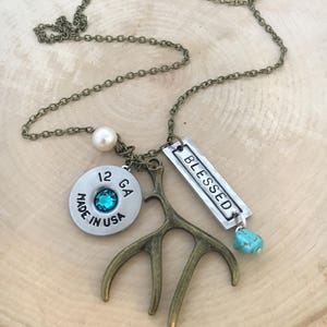 May include: A silver necklace with a 12 GA shotgun shell charm, a turquoise stone, a small pearl, and a silver charm that says "Blessed". The necklace also features a bronze antler charm.