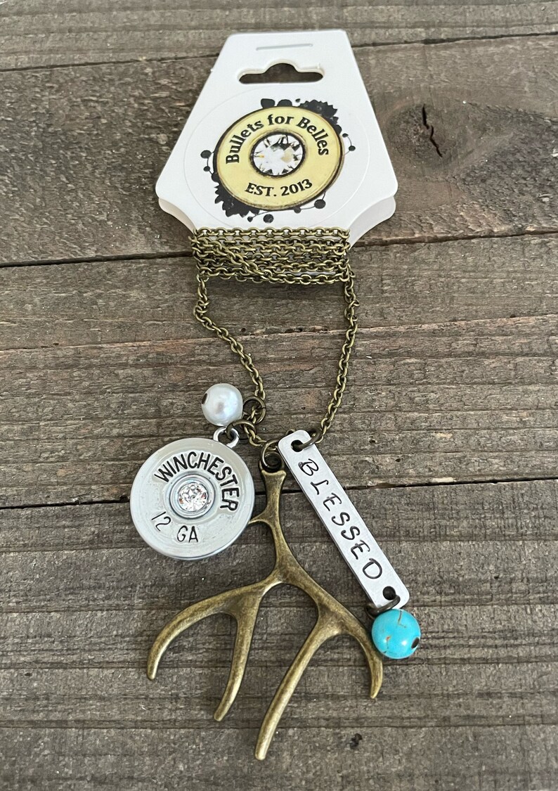 May include: A necklace with a silver bullet charm that says "Winchester 12 GA", a small antler charm, a silver charm that says "Blessed", a white pearl bead, and a turquoise bead. The necklace is on a brown wooden surface.