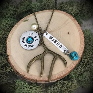 May include: A necklace with a 12 GA shotgun shell, a turquoise stone, a small antler charm, a pearl bead, and a silver charm that says "BLESSED".