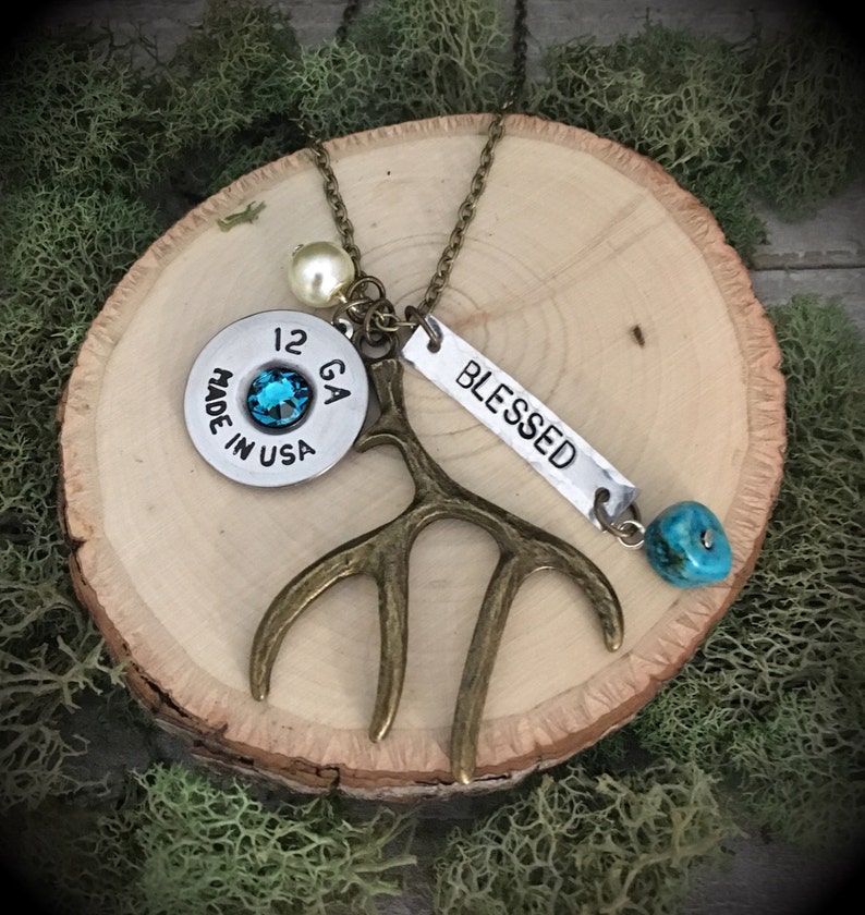 May include: A necklace with a 12 GA shotgun shell, a turquoise stone, a small antler charm, a pearl bead, and a silver charm that says "BLESSED".