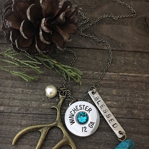 May include: A silver chain necklace with multiple charms, including a silver charm with the words "Winchester 12 GA" and a turquoise stone. The necklace also features a small pearl, a silver charm with the word "Blessed" and a pair of bronze antlers.