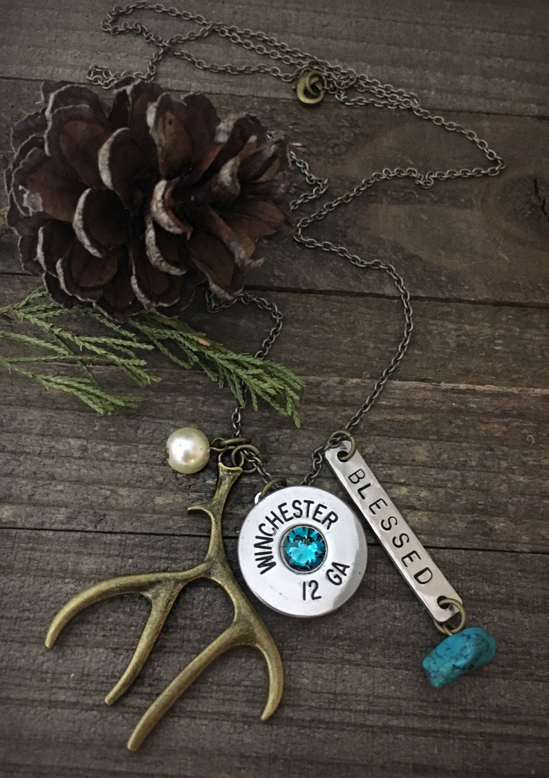 May include: A silver chain necklace with multiple charms, including a silver charm with the words "Winchester 12 GA" and a turquoise stone. The necklace also features a small pearl, a silver charm with the word "Blessed" and a pair of bronze antlers.