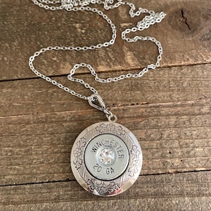 May include: Silver-toned pendant necklace featuring a Winchester 20 GA bullet casing with a central clear crystal. The pendant is round, with floral detailing around the casing. The chain is a delicate silver link.