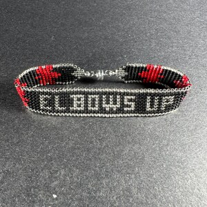 Puede incluir: Pulsera de cuentas negras y rojas con cierre plateado. La pulsera tiene las palabras "BOWS OUT" en cuentas blancas.