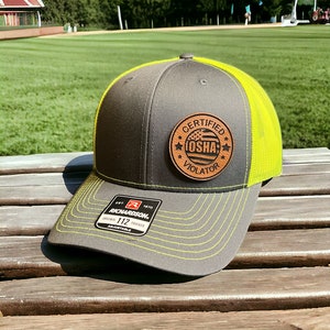 May include: A gray and neon yellow trucker hat with a brown leather patch that reads "Certified OSHA Violator" with a small American flag inside the circle. The hat is made by Richardson 112.