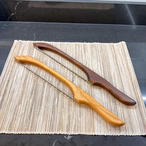 May include: Two wooden cheese slicers with curved handles, one crafted from light-toned wood and the other from dark-toned wood. Each slicer features a thin, taut metal wire stretched between the ends of the handle for cutting through cheese.