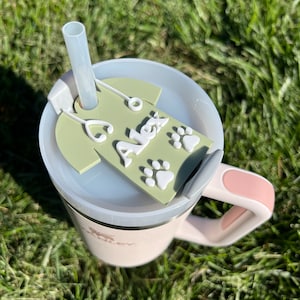 May include: A white travel mug with a pink handle and a green personalized tag that says "Alex" with paw prints. The mug has a clear lid with a straw.