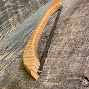 May include: A wooden-handled saw with a curved blade. The handle is made of maple wood with a natural grain pattern. The blade is made of metal and has a serrated edge.