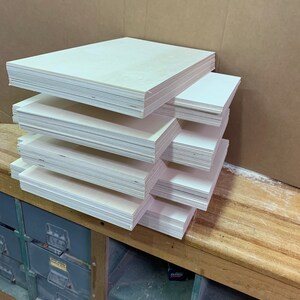 Puede incluir: Varias pilas de paneles de madera rectangulares de color claro est&aacute;n dispuestas sobre un banco de trabajo de madera. Los paneles est&aacute;n cuidadosamente apilados en varias pilas, mostrando los bordes de la madera. El fondo incluye una caja de cart&oacute;n y un armario met&aacute;lico.