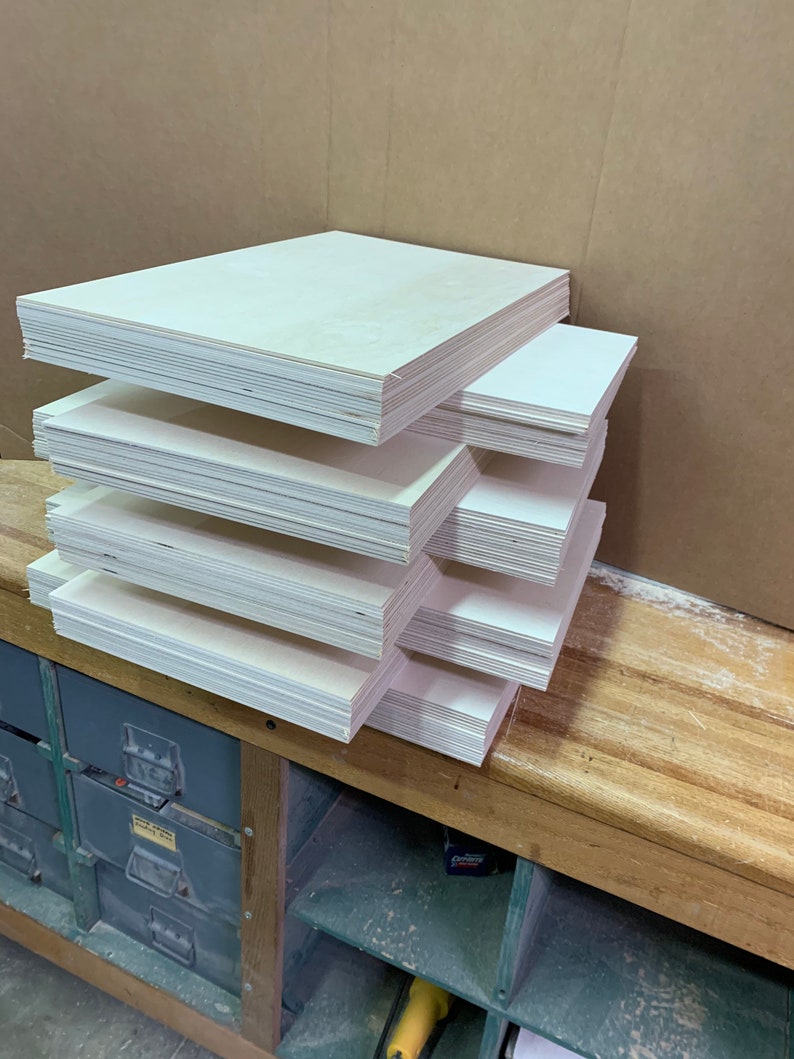 Puede incluir: Varias pilas de paneles de madera rectangulares de color claro est&aacute;n dispuestas sobre un banco de trabajo de madera. Los paneles est&aacute;n cuidadosamente apilados en varias pilas, mostrando los bordes de la madera. El fondo incluye una caja de cart&oacute;n y un armario met&aacute;lico.