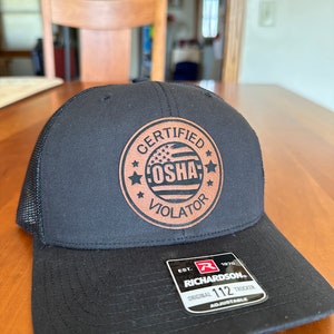 May include: Black trucker hat with a mesh back and a curved brim. The hat features a brown leather patch with the words "CERTIFIED OSHA VIOLATOR" and a US flag design. The hat also has a Richardson 112 Trucker tag.