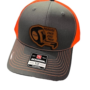 May include: A gray and orange trucker hat with a brown leather patch featuring a tape measure and the text "MEASURE ONCE CUSS TWICE". The hat is adjustable and has the Richardson logo and the text "ORIGINAL 112 TRUCKER ADJUSTABLE".