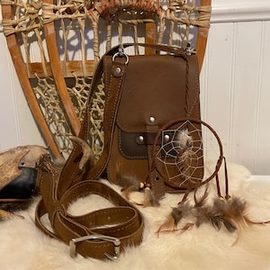 May include: A brown leather crossbody bag with a flap closure, a matching belt, and a small dreamcatcher with feathers. A vintage snowshoe is in the background. The items are displayed on a white, fluffy surface.