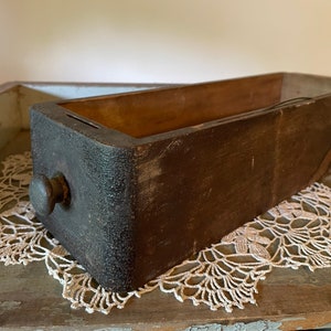 May include: A weathered, rectangular wooden drawer with a dark brown finish and a round knob. The drawer's interior is a lighter shade of brown. The drawer is resting on a white lace doily.