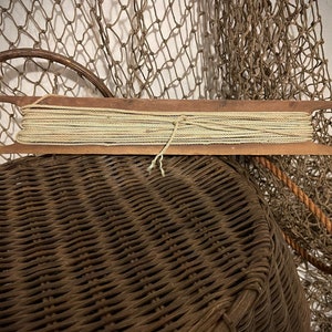 May include: A vintage wooden spool filled with beige twine, resting on a woven wicker basket. The spool has a rectangular shape with pointed ends. A fishing net hangs in the background, adding to the rustic, nautical theme.