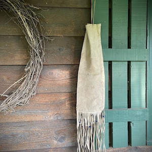May include: A tan suede pouch with long fringe and blue beads hanging against a weathered wooden background. The pouch is suspended by a thin cord. A rustic wreath is visible on the left side of the image.