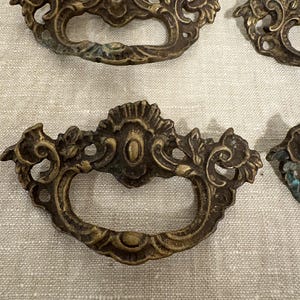 May include: Ornate antique brass drawer pulls with a floral design. The pulls have a curved handle and decorative detailing. The brass has a weathered appearance with hints of green patina, suggesting age and use. These are likely vintage or antique cabinet hardware.