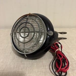 May include: A vintage black headlight with a clear glass lens and a protective metal grill. The headlight has a mounting bracket and red and black wires. The design is reminiscent of classic automotive or industrial lighting.