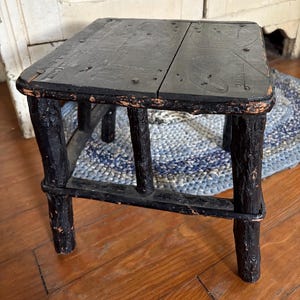 May include: A small, rectangular, black wooden stool with four legs. The stool has a rustic, distressed appearance, with visible wood grain and nail heads. The legs are connected by horizontal supports.