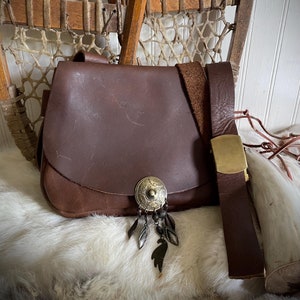 May include: A brown leather satchel with a flap closure, adorned with a decorative metal emblem and dangling charms. The satchel has a matching leather strap with a brass buckle. The bag is displayed on a white fur-like surface.