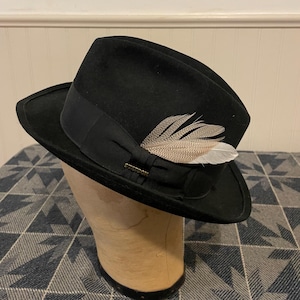 May include: A black fedora hat with a black band and a decorative feather. The hat is displayed on a mannequin head. The hat has a brim and a small metal tag that reads "Stetson".